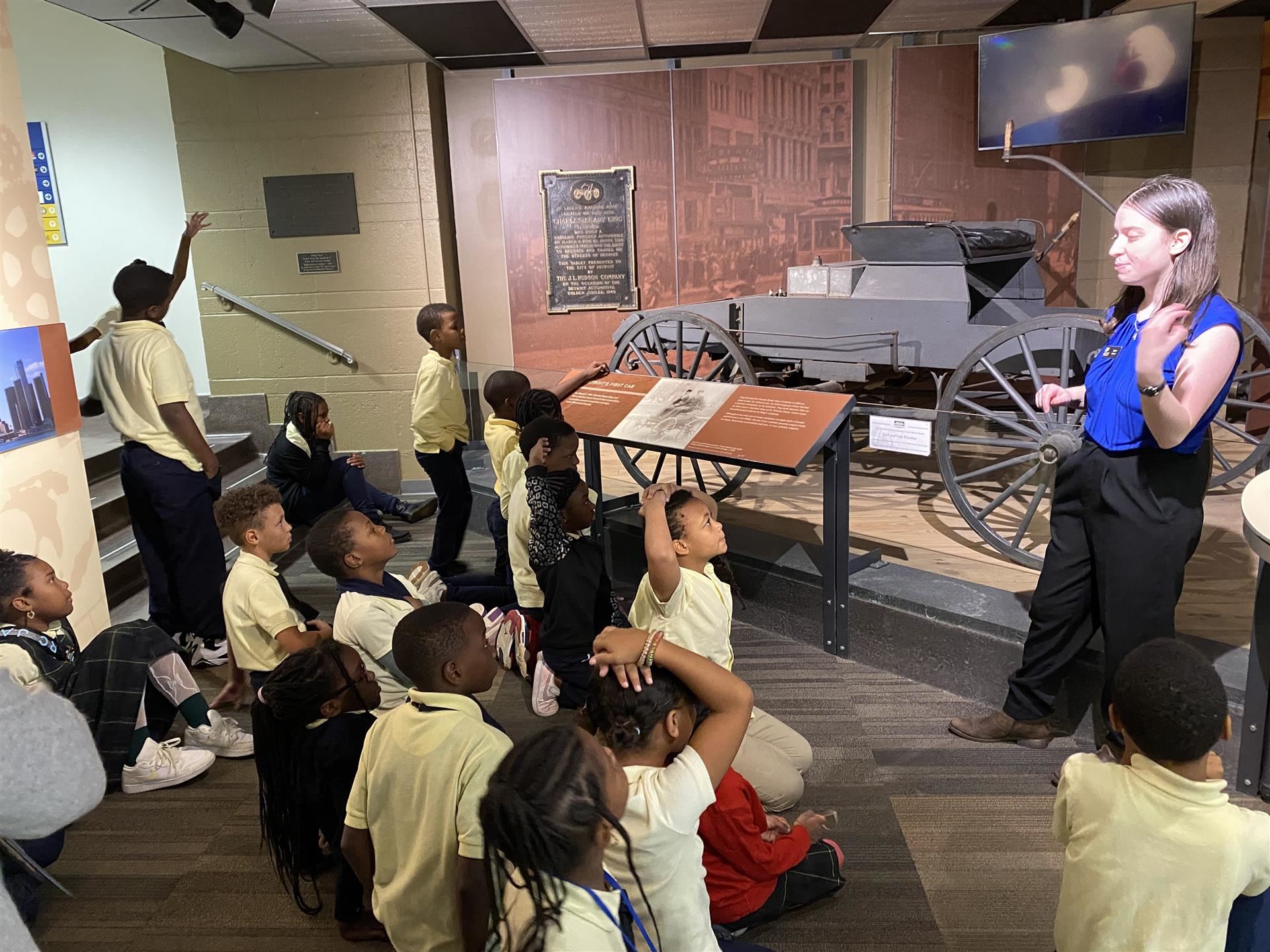 class of students sitting on the floor looking at tour guide and old car